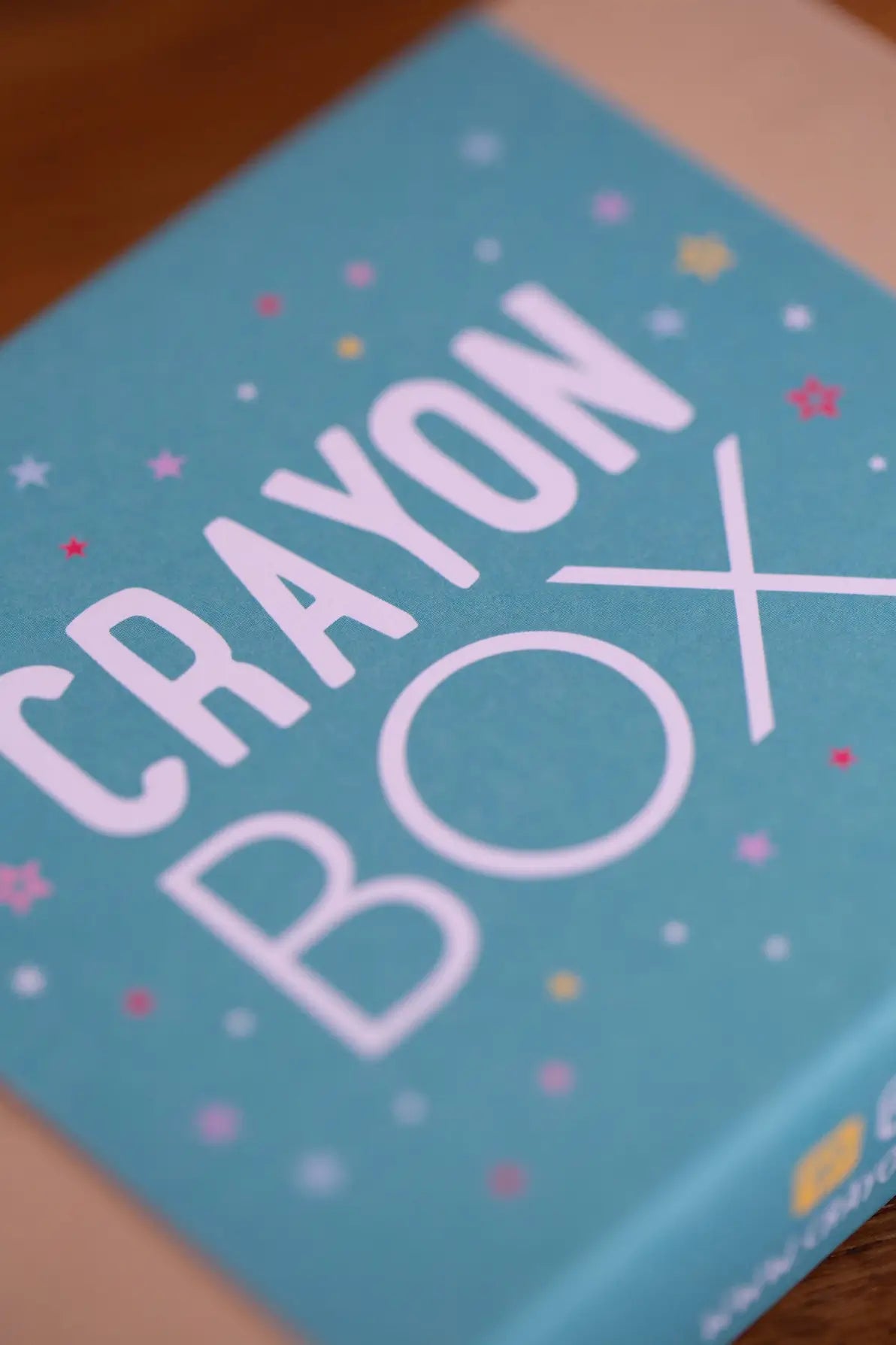 A close-up angled shot of a light blue box with large white text reading 'CRAYON BOX' and small pastel star and dot illustrations on the cover, resting on a wooden surface.