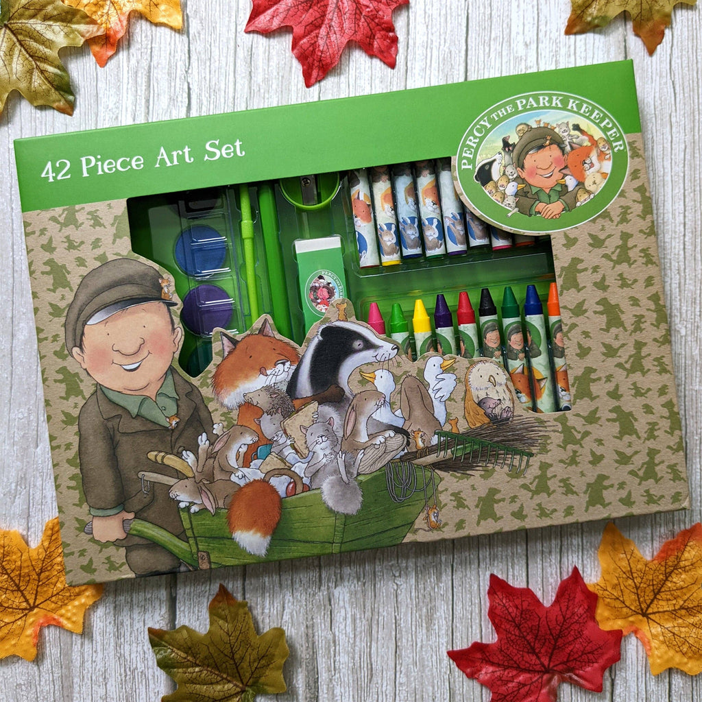 A 42-piece art set box for 'Percy the Park Keeper' displayed on a rustic wood background with autumn leaves. The box features woodland illustrations of Percy and various animals in a cart, with crayons, markers, and art supplies visible through a clear window.