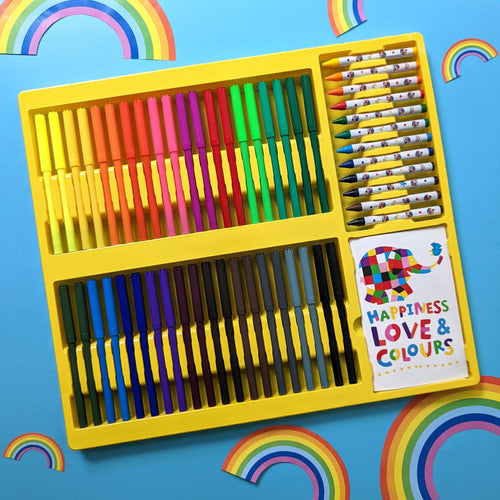 A children's art set in a bright yellow plastic tray, featuring rows of colorful markers and crayons, arranged on a blue background decorated with rainbow cutouts. A small card on the tray reads 'HAPPINESS LOVE & COLOURS' with a colorful elephant illustration.