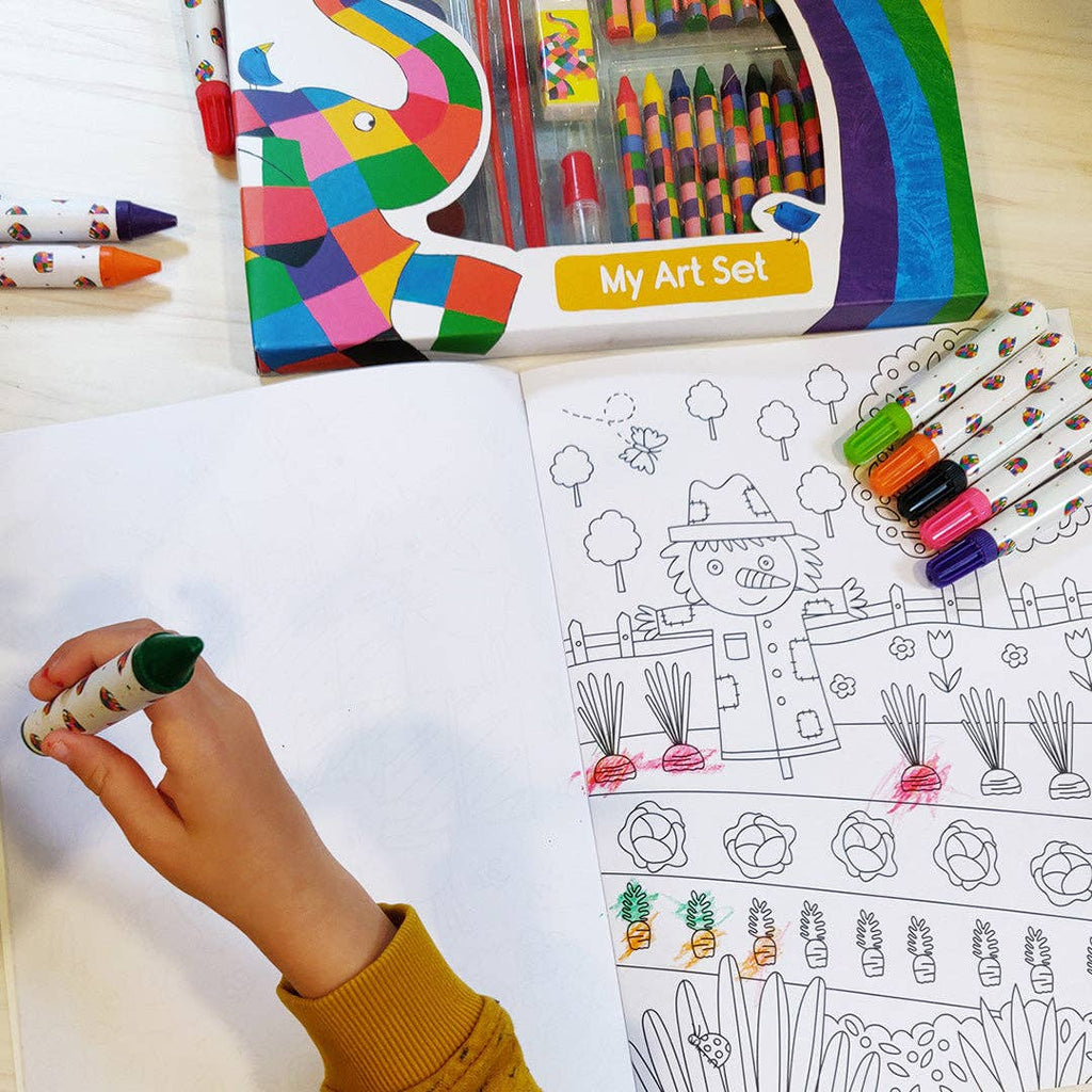 A children's art set shown in use: an open colouring book with black-and-white garden illustrations, a hand holding a green marker, several crayons, and a box labelled 'My Art Set' on a light wood surface.