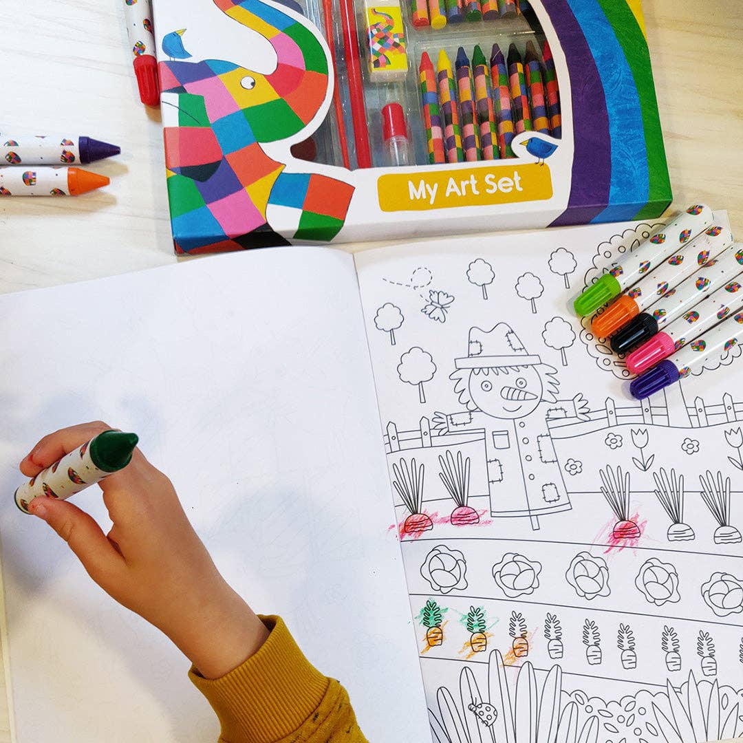 A children's art set shown in use: an open colouring book with black-and-white garden illustrations, a hand holding a green marker, several crayons, and a box labelled 'My Art Set' on a light wood surface.