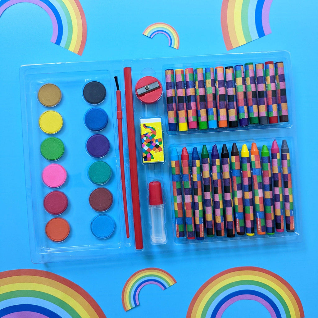 A children's art and craft kit in a clear plastic case, featuring crayons, watercolour paints, a paintbrush, a small red pencil sharpener, a matchbox, and a small clear bottle with red cap, arranged on a bright blue background with rainbow decorations.