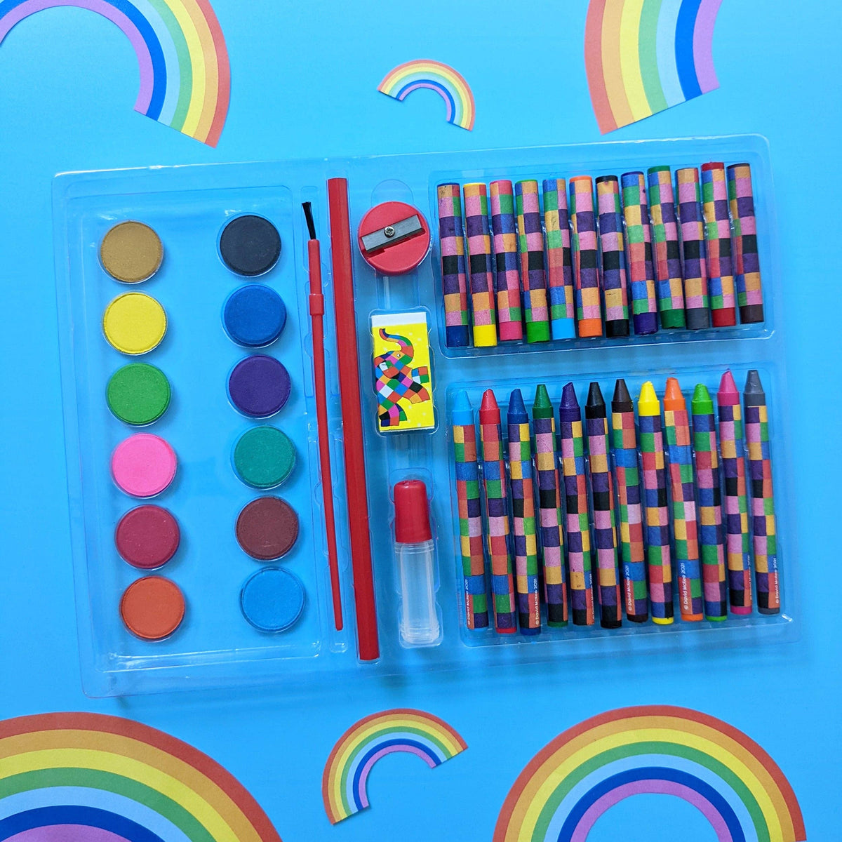 A children's art and craft kit in a clear plastic case, featuring crayons, watercolour paints, a paintbrush, a small red pencil sharpener, a matchbox, and a small clear bottle with red cap, arranged on a bright blue background with rainbow decorations.