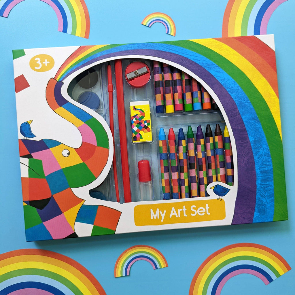 A children's art set in a colourful box with rainbow graphics and an elephant illustration. The set includes crayons, coloured pencils, a small paintbrush, a sharpener, and a small paint palette, shown on a bright blue background with paper rainbow decorations.