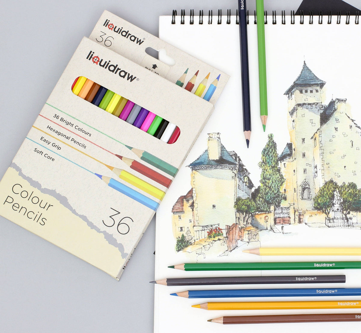 A 36-piece set of liquidraw hexagonal colour pencils in a beige cardboard box, styled on a gray surface next to a spiral sketchbook showing a watercolour drawing of a castle. Several coloured pencils are placed on top of the sketchbook.
