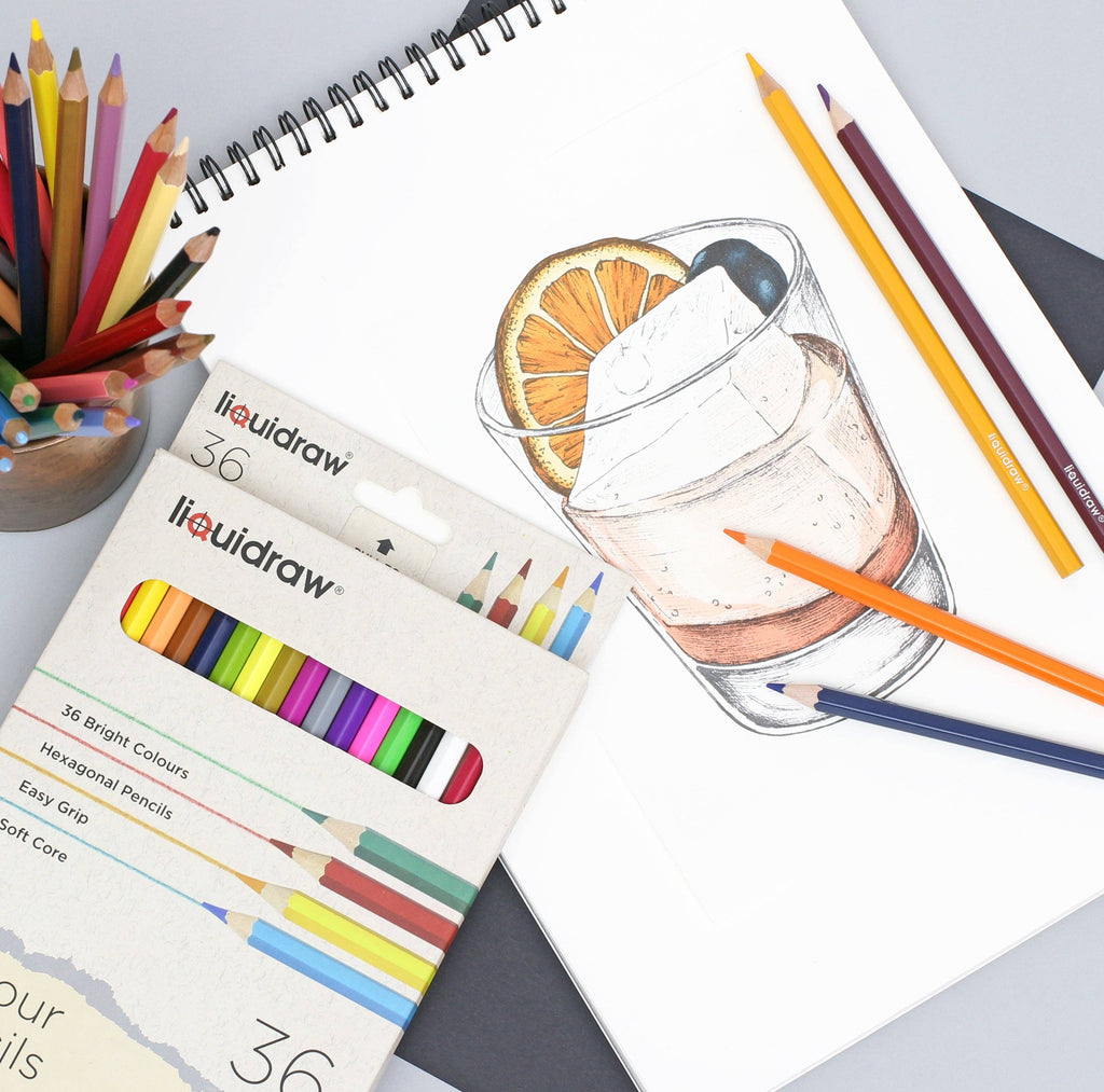 A Liquidraw 36 pastel pencil set in its original packaging, styled on a drawing pad next to a sketch of a cocktail and surrounded by colored pencils.