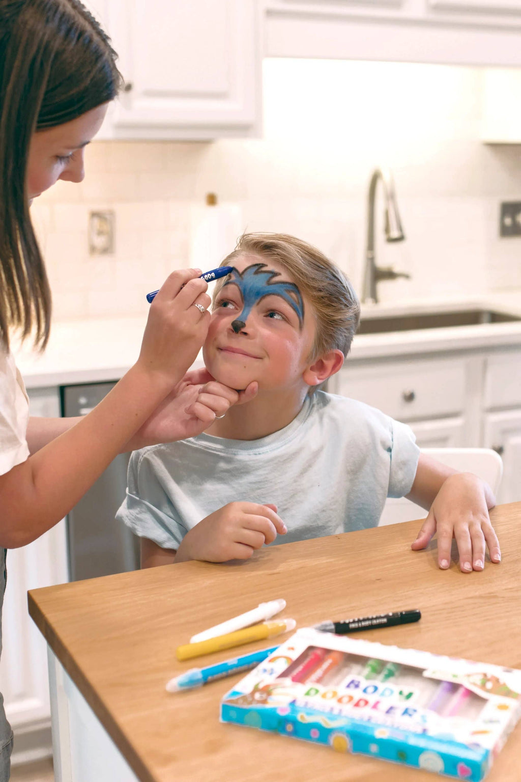 Person applying face paint to a child in a kitchen setting