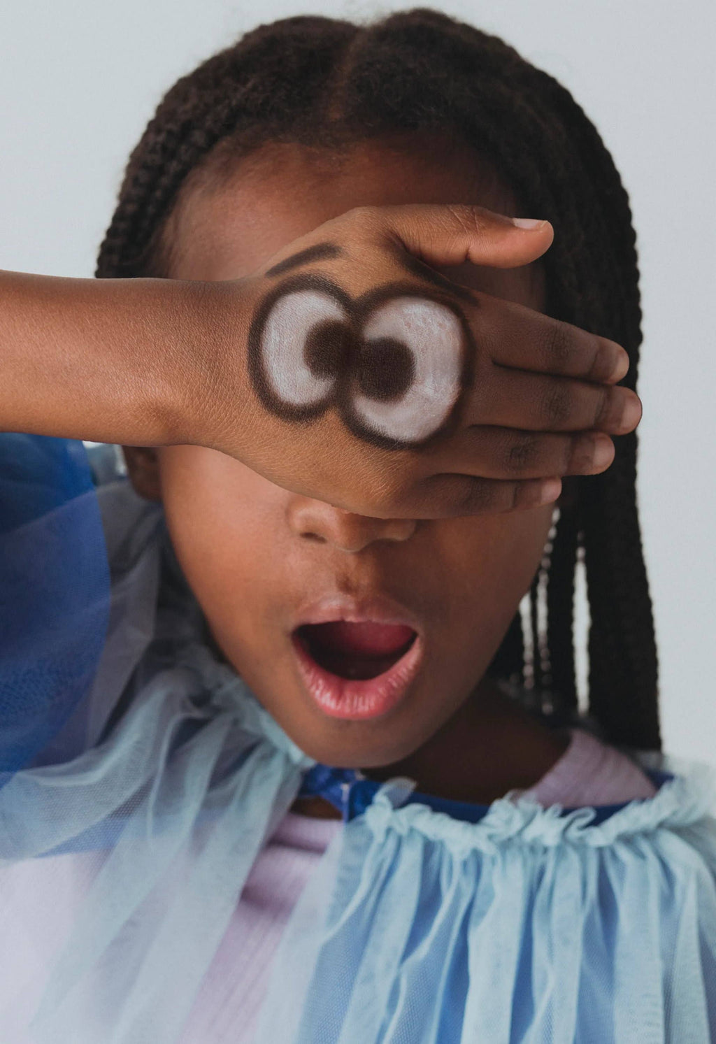 Child with braided hair wearing a blue dress, covering their eyes with a hand.