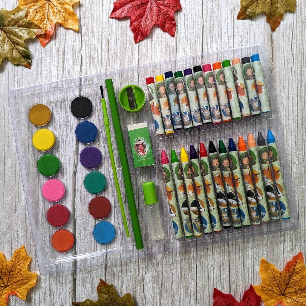 A children's art and craft set arranged in a clear plastic case on a whitewashed wood surface with autumn leaves around the edges. The set includes a round palette of colorful paint pans, two green pencils, a green eraser, a small brush, crayons, markers, and other drawing tools.