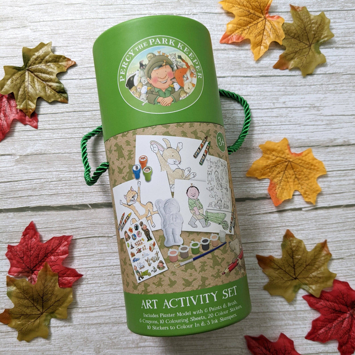 A cylindrical green and brown cardboard art activity set tube labelled 'Percy the Park Keeper Art Activity Set,' surrounded by scattered autumn leaves on a whitewashed wood surface.