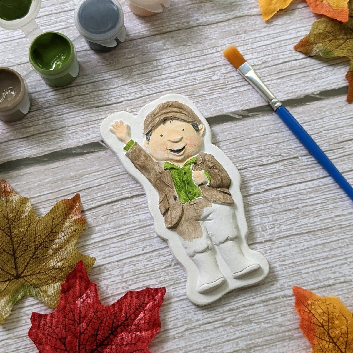 A hand-painted ceramic figure of a smiling Percy the Park Keeper in an autumn outfit, surrounded by paint pots, a paintbrush, and colourful fall leaves on a light wood background.