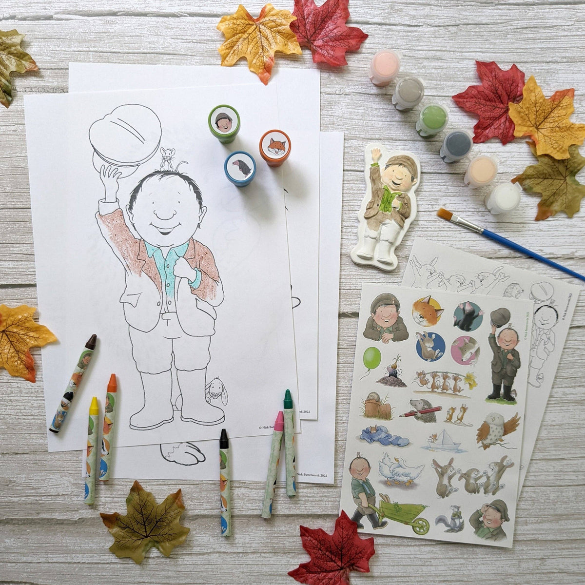 A styled flat-lay of a children's colouring and sticker activity set on a whitewashed wood background, surrounded by autumn leaves. The set includes a black-and-white colouring page of a boy holding a football, a sticker sheet with cartoon characters, a paint palette, crayons, and paint pots.