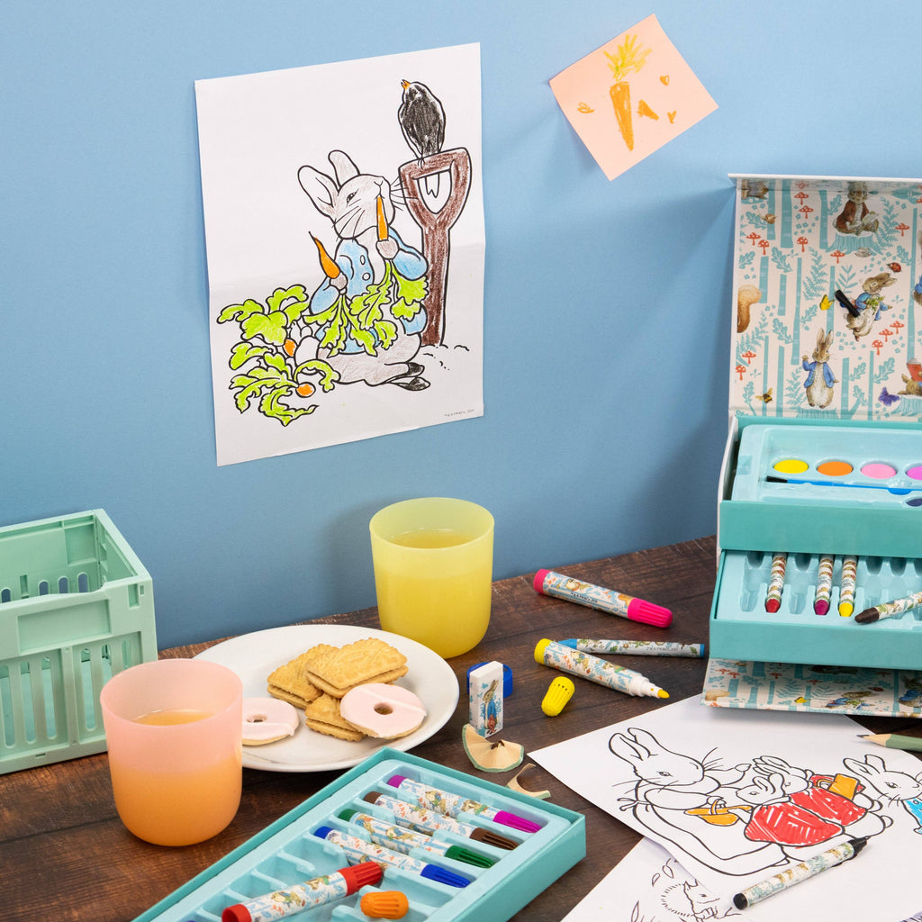 A children's art and colouring set arranged on a wooden table against a blue wall, including a turquoise box with watercolour palettes, markers, crayons, and printed colouring pages featuring a rabbit and carrot illustrations.
