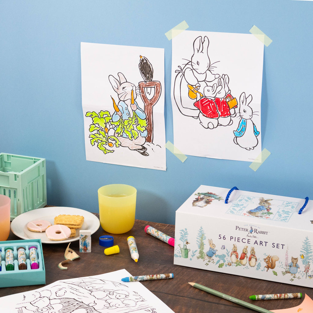 A children's art set displayed on a wooden table against a light blue wall. The set includes a white box labelled 'Peter Rabbit 56 Piece Art Set,' coloured pastels in a teal case, crayons, drawing pencils, a yellow cup, cookies on a plate, and two printed rabbit colouring pages taped to the wall.