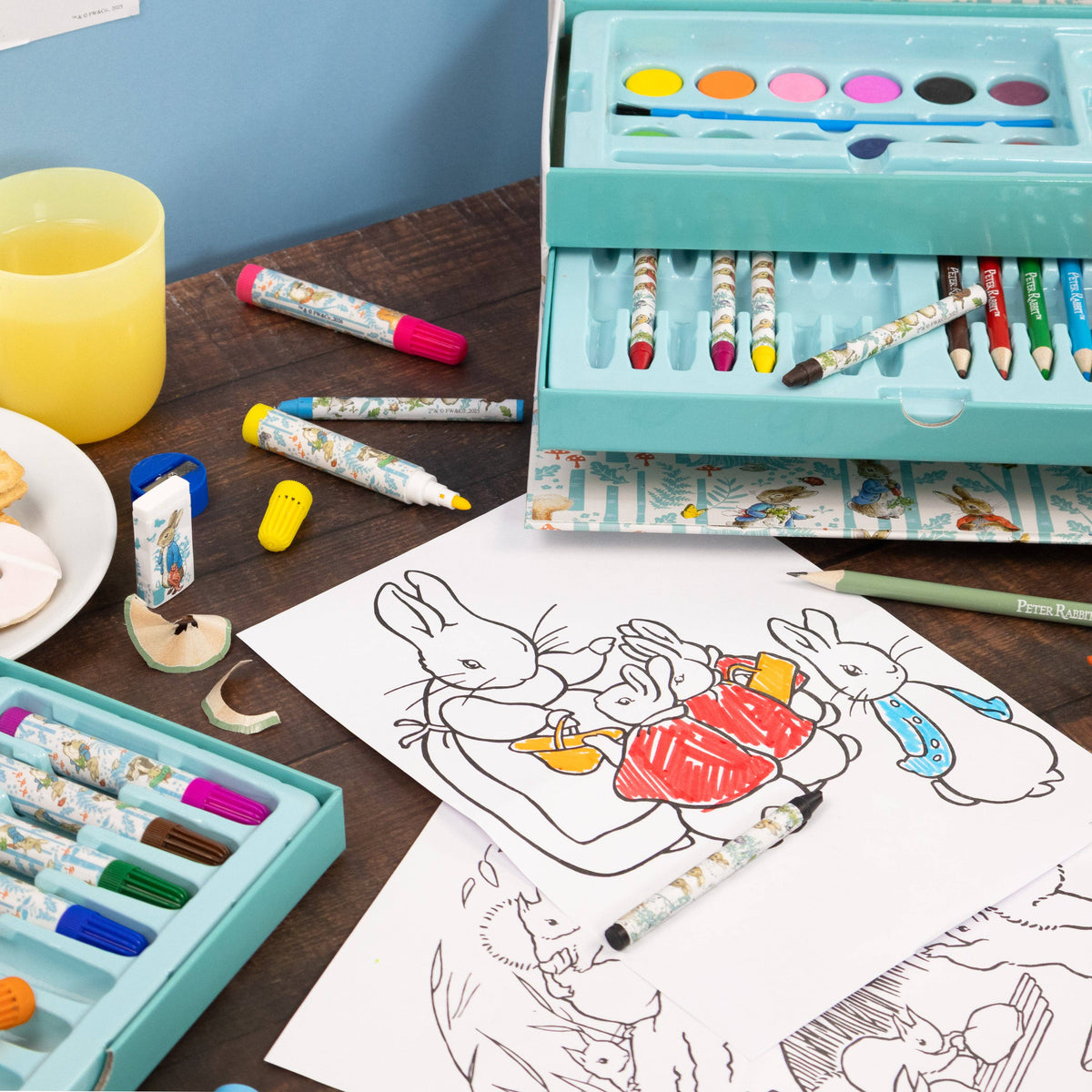 A children's art set displayed on a wooden table with colouring pages, crayons, markers, a watercolour palette, and a pencil. The set includes a turquoise storage case with patterned crayons, a matching marker set, and a watercolour palette, alongside loose art supplies.