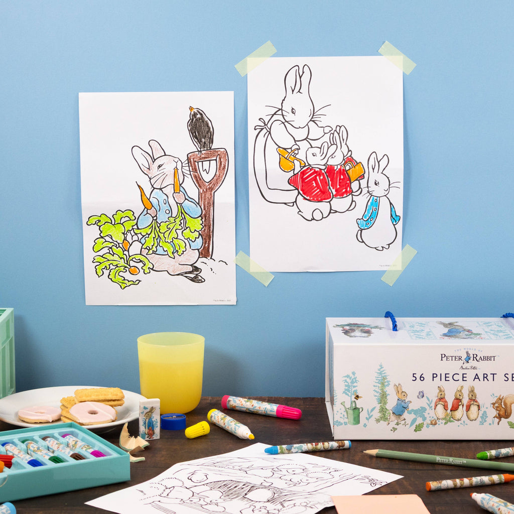 Two Peter Rabbit colouring pages taped to a light blue wall above a desk with art supplies, including a box labelled Peter Rabbit 56 Piece Art Set.
