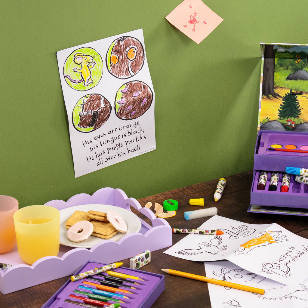 A children's colouring activity setup on a wooden table against a green wall, featuring a purple crayon box, colouring pages, a tray with donuts and a drink, and a printed colouring book page pinned on the wall.