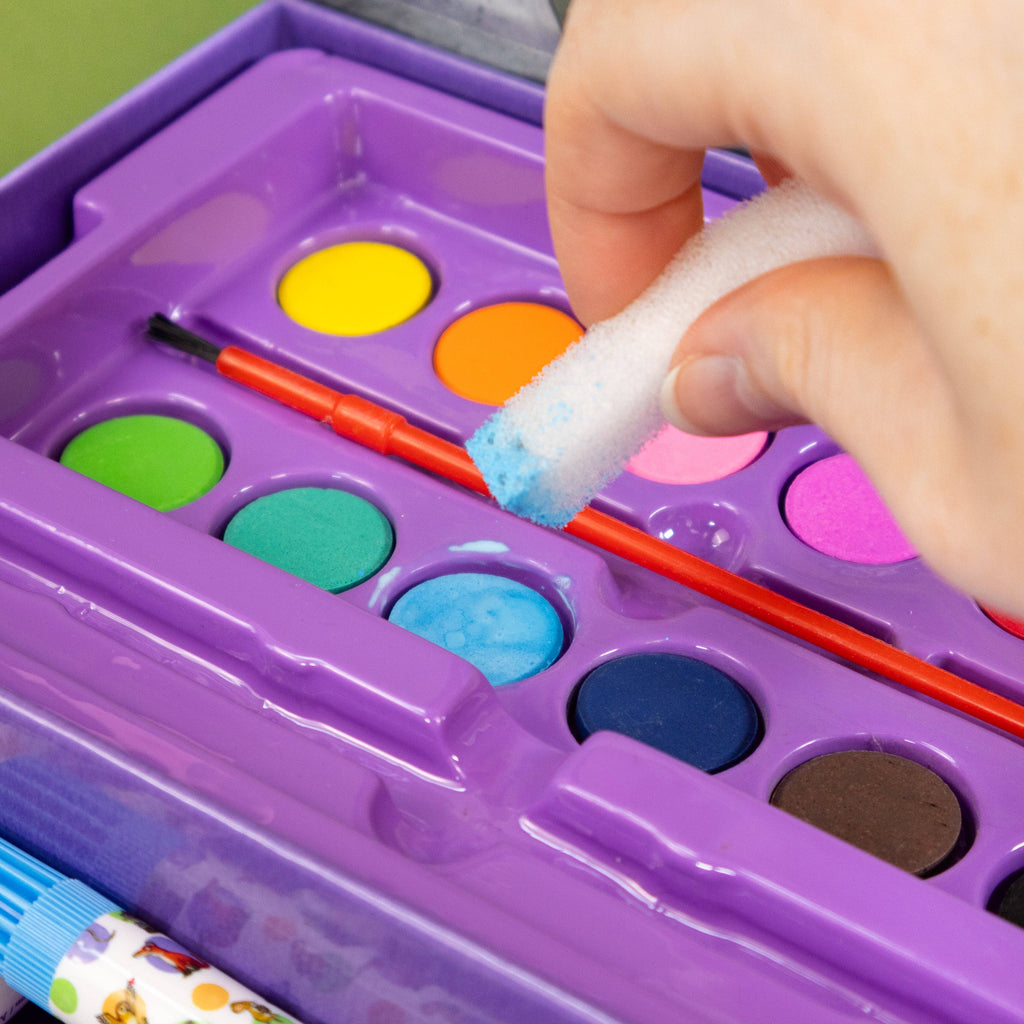 A close-up of a purple watercolour paint palette with colourful paint wells, a small orange paintbrush, and a hand holding a white foam sponge dipped in blue paint, with part of a glue tube visible at the bottom left.