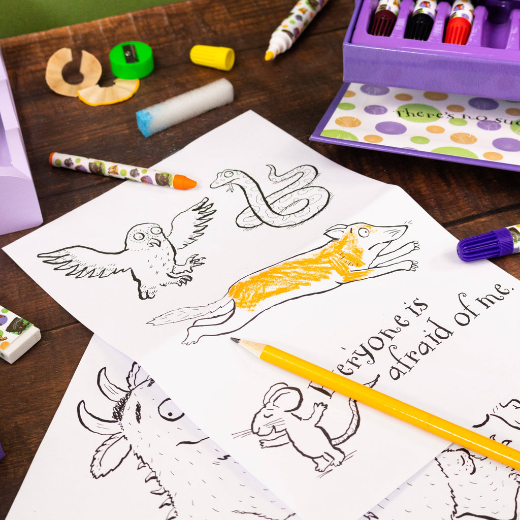 A styled flat-lay of children's colouring pages on a wooden table, surrounded by colourful crayons, a purple marker tray, and a yellow pencil. The main page shows black-and-white line drawings of an owl, a snake, a dog with one leg raised, a mouse, and the phrase 'everyone is afraid of me' in script.
