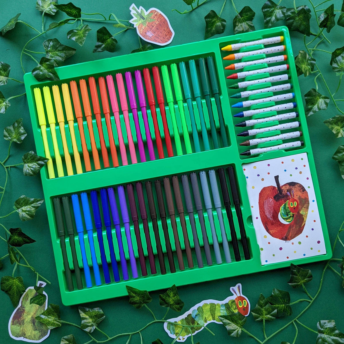 Colourful art supply set with markers, crayons, and pencils on a green background with leaf decorations.