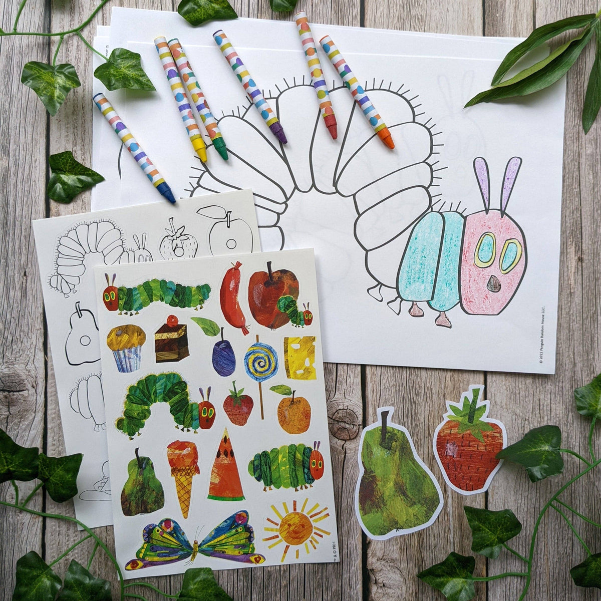 Colouring sheets and stickers of a caterpillar and fruits on a wooden surface with leaves.