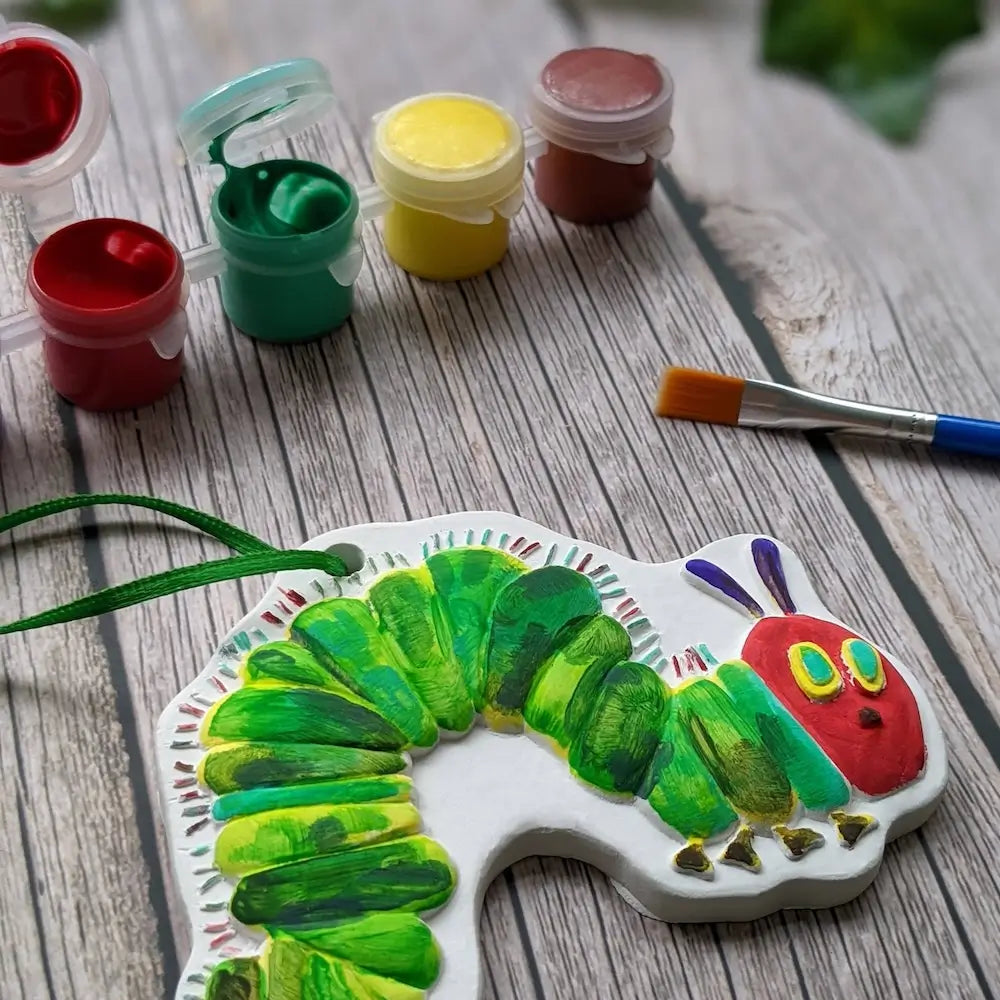 Painted wooden cutout of a caterpillar with paint containers and a brush on a wooden surface.
