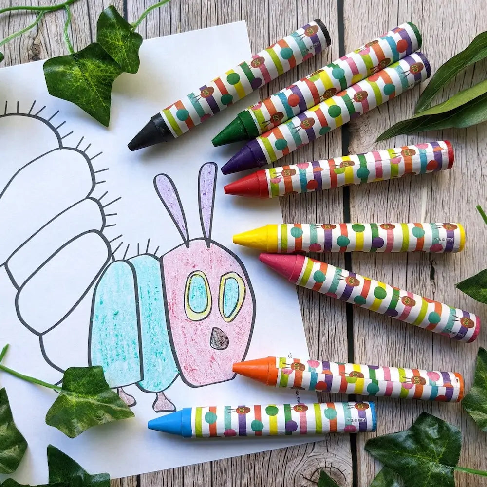 A set of colorful crayons with striped paper-wrapped handles arranged on a wooden surface next to a coloring page of a caterpillar, with green ivy leaves scattered around.
